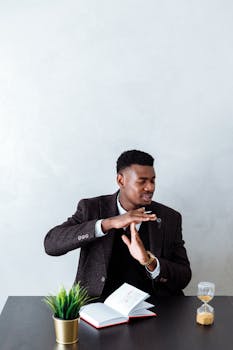 Businessman in a black blazer signaling time out with an hourglass and open book on a table.