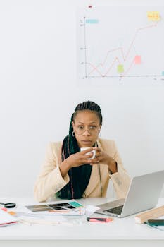 Focused businesswoman holding cup, analyzing financial data at desk.