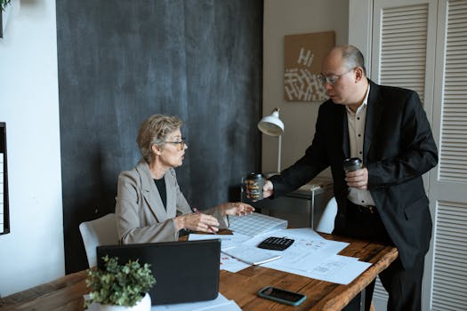 Two professionals in a modern office discussing work over coffee. Papers and laptop on desk.